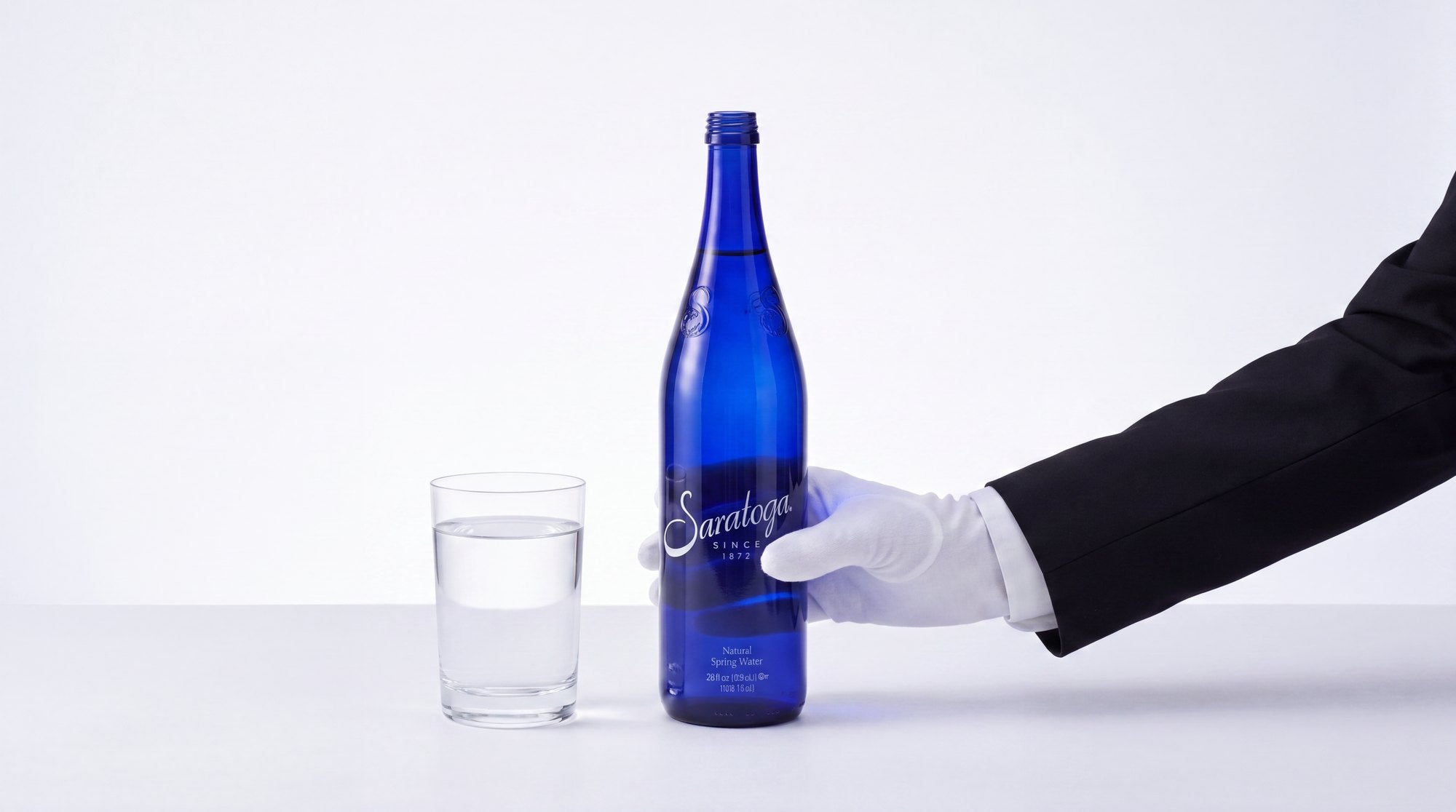 A white-gloved hand places a blue Saratoga bottle beside a glass of water